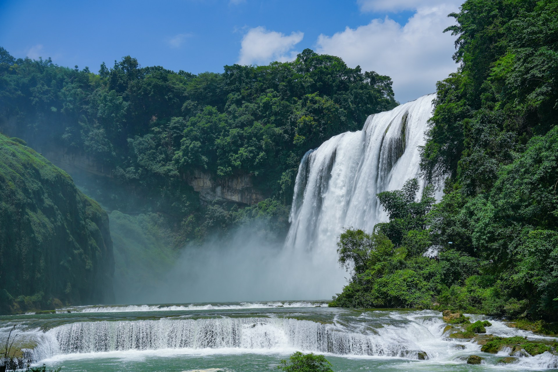 A panoramic view of Huangguoshu Waterfall Scenic Area in Guizhou, China, showing Asia's largest waterfall cascading into Rhinoceros Pool surrounded by lush karst valley vegetation.