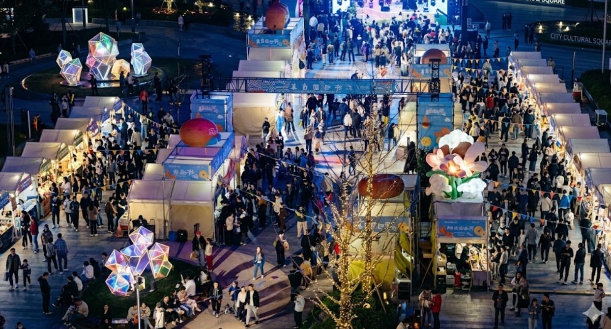 Aerial night view of Shanghai 55 Shopping Festival outdoor market with colorful light installations, crowded vendor stalls, and large decorative sculptures at City Cultural Square