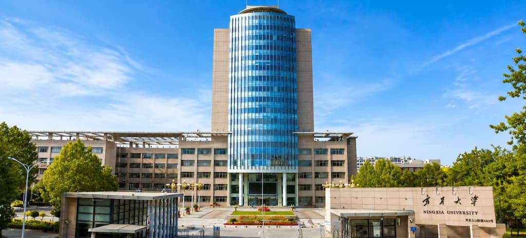 Wide-angle daytime view of Ningxia University campus main building with central glass tower, landscaped plazas, and surrounding greenery under a clear blue sky.