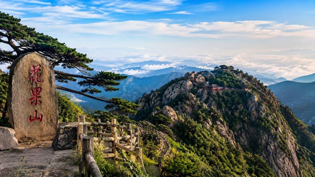Panoramic view of Huaguoshan Scenic Area featuring rocky mountain peaks, lush greenery, traditional Chinese buildings, and a stone monument overlooking a sea of clouds.