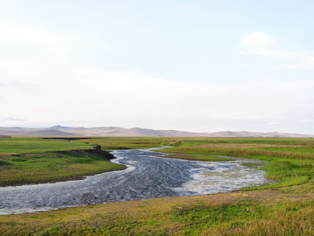 Panoramic view of Hulunbuir grasslands with a winding river, lush green fields, and distant rolling hills under a clear sky.