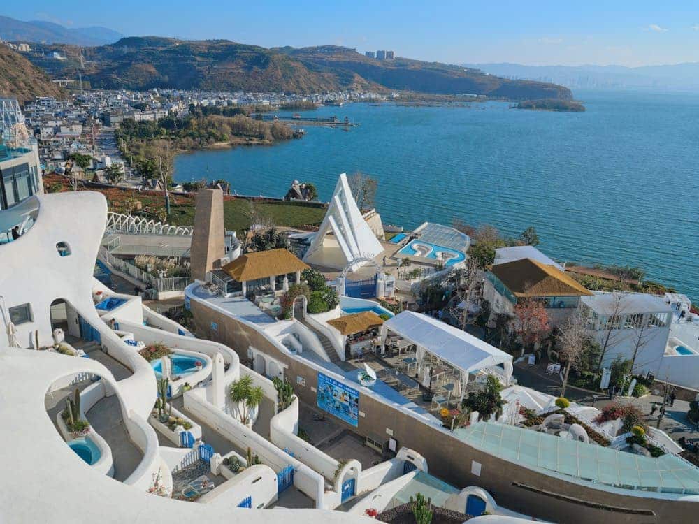 A panoramic coastal view of Dali beside Erhai Lake featuring white Santorini-style architecture, hillside villages, and clear blue water under sunny skies.