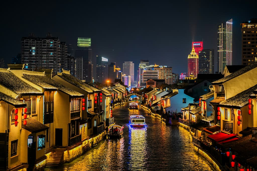 A wide-angle, serene view of Jichang Garden in Liangxi District, Wuxi, showing a zigzag wooden bridge over a lotus pond with traditional Chinese pavilions and ancient trees.