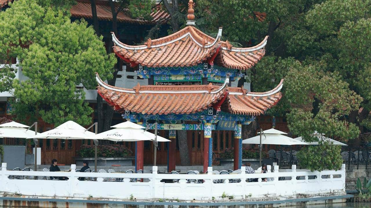 Traditional Chinese wooden pavilion beside a calm lake with lush green trees reflected in still water, Beijing imperial garden.