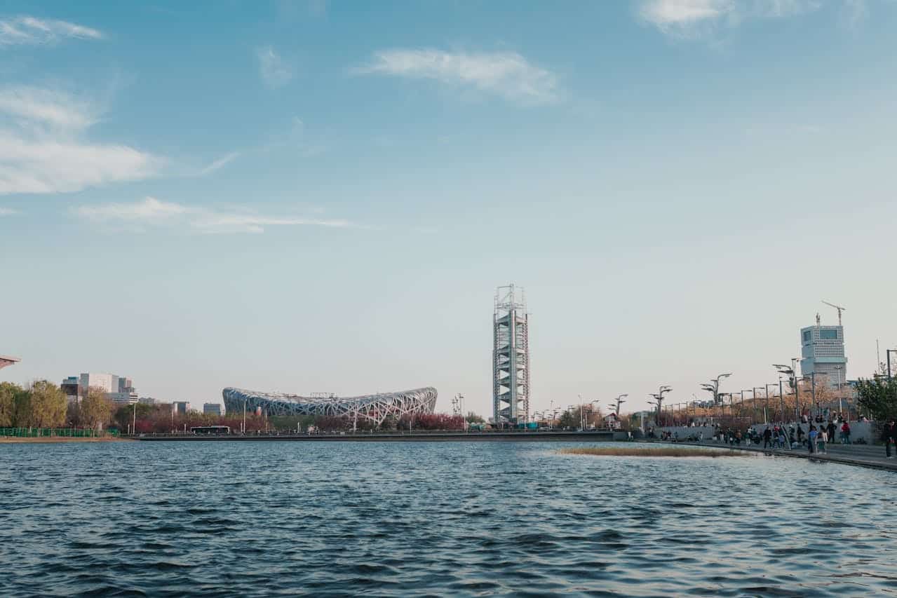 Panoramic view of Beijing Olympic Park with water in the foreground, showing the iconic Bird’s Nest Stadium and nearby observation tower under a clear sky, with trees and visitors along the park pathways.