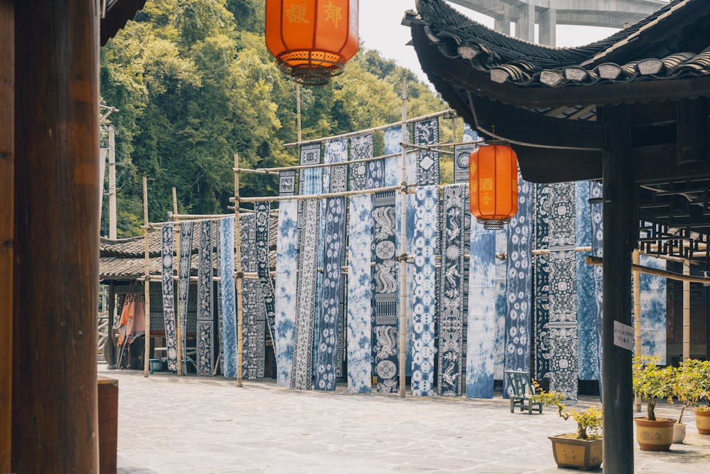 Long panels of blue-and-white indigo tie-dye fabric hanging to dry on bamboo frames in a traditional Chinese courtyard with red lanterns