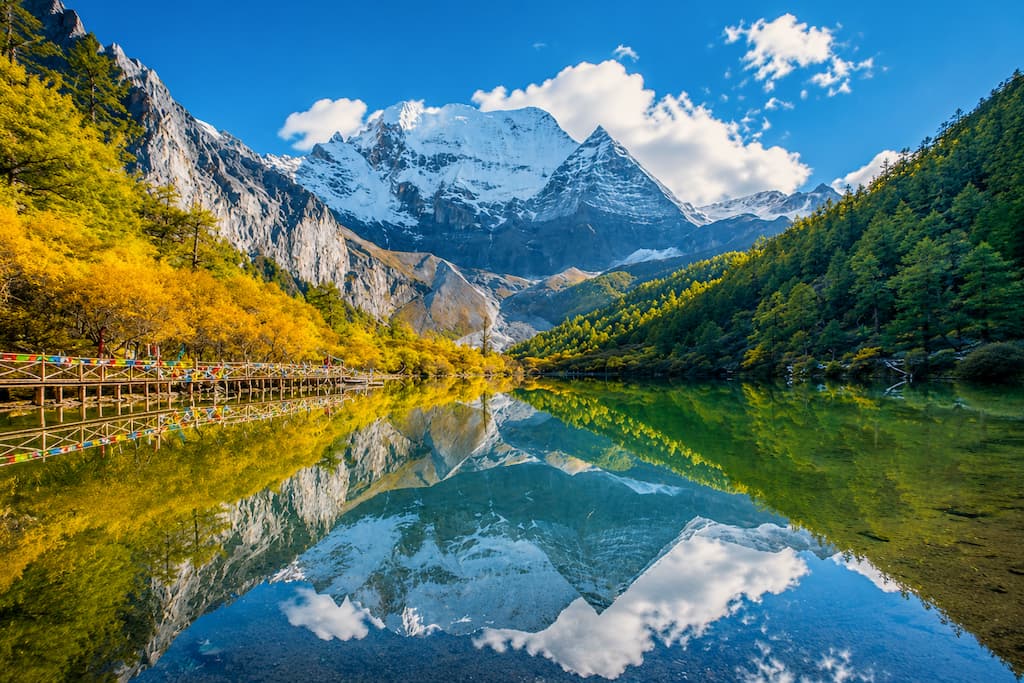 Snow-capped peaks of Daocheng Yading reflected in a turquoise alpine lake framed by golden autumn larches and Tibetan prayer flags