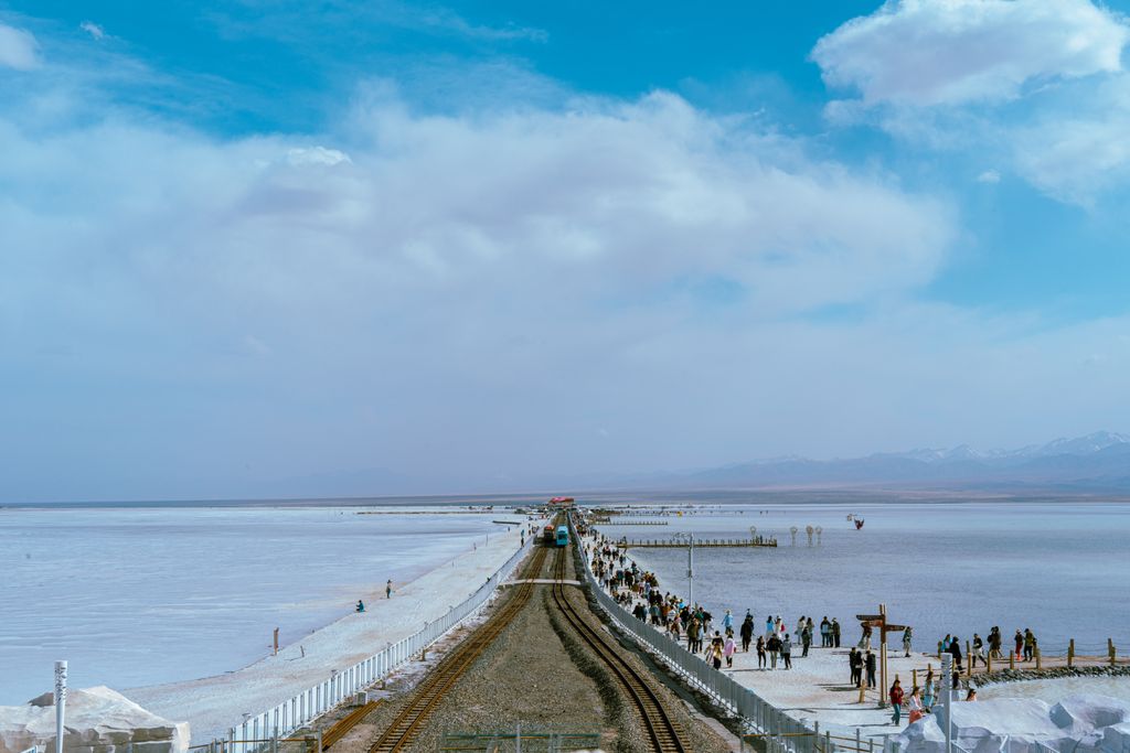 Sightseeing train and tourists on the railway crossing the white salt flats of Chaka Salt Lake, with snow-capped mountains in the distance