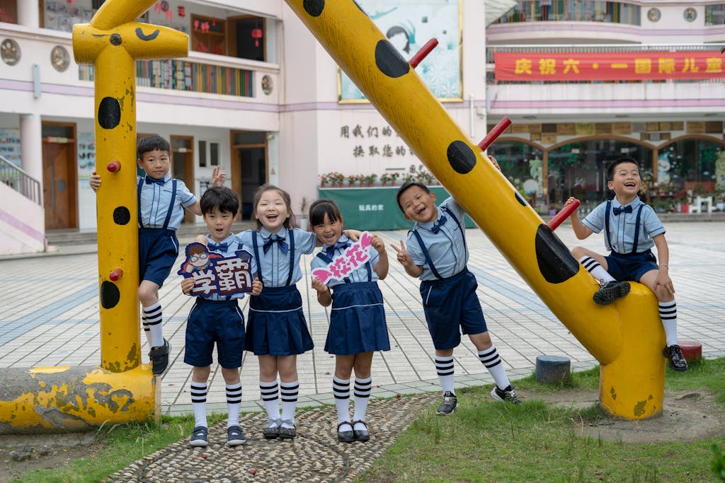 A group of joyful Chinese school children wearing matching blue and white uniforms pose playfully around a yellow giraffe-shaped structure in a school courtyard, holding colorful cartoon signs during a Children's Day celebration.